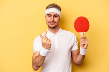Young caucasian man holding ping pong rackets isolated on yellow background pointing with finger at you as if inviting come closer.