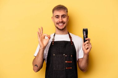Young caucasian hairdresser man isolated on yellow background cheerful and confident showing ok gesture.