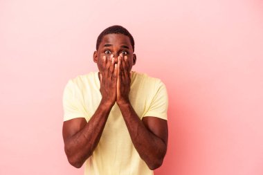 Genç Afrikalı Amerikalı bir adam pembe arka planda izole edilmiş. Şok olmuş, ağzı ellerle kaplanmış, yeni bir şey keşfetmeye hevesli..