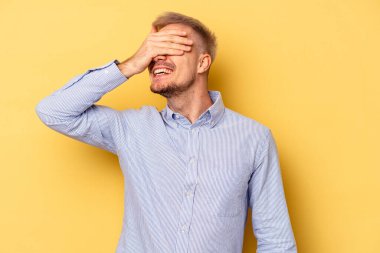 Sarı arka planda izole edilmiş genç beyaz adam elleriyle gözlerini kapatıyor, geniş bir gülümsemeyle bir sürpriz bekliyor..