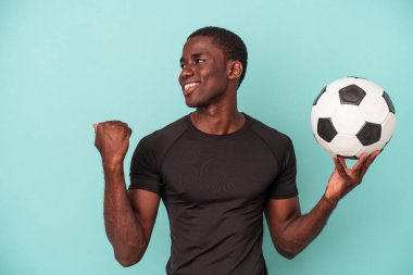 Futbol oynayan genç Afrikalı Amerikalı adam mavi arka planda izole edilmiş, başparmak uzakta, gülüyor ve kaygısız..