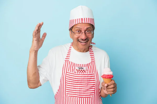 Senior american ice cream man holding an ice cream isolated on blue background receiving a pleasant surprise, excited and raising hands.