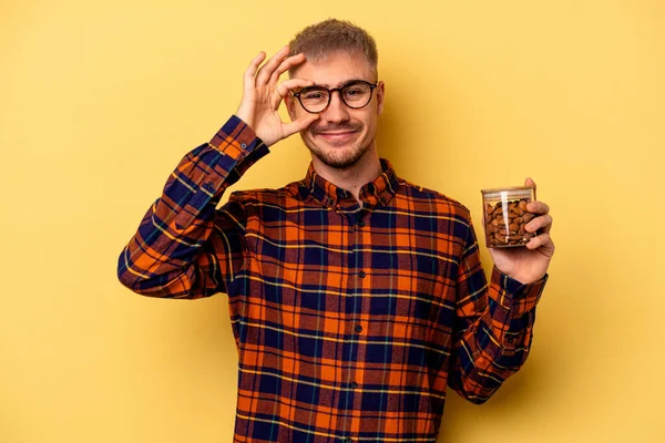 Young caucasian man holding a almond jar isolated on yellow background excited keeping ok gesture on eye.