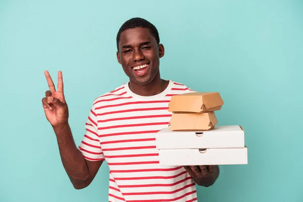 Молодой Афроамериканец Держит Пиццу Бургеры Голубом Фоне Радуясь Беззаботно Показывая — стоковое фото