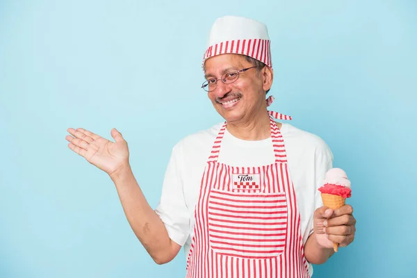 Senior american ice cream man holding an ice cream isolated on blue background showing a copy space on a palm and holding another hand on waist.