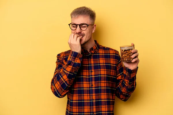 Young Caucasian Man Holding Almond Jar Isolated Yellow Background Biting — Zdjęcie stockowe