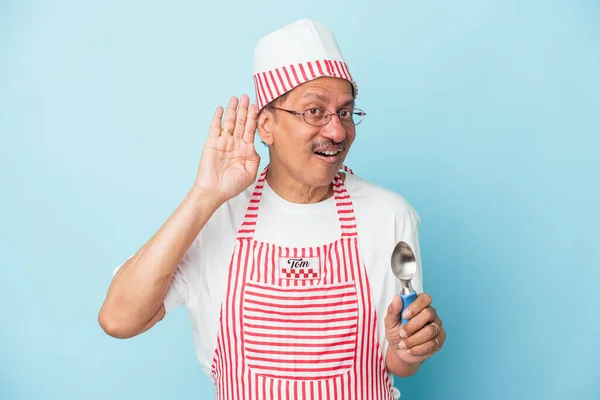 Senior indian ice cream man holding a scoop isolated on blue background trying to listening a gossip.