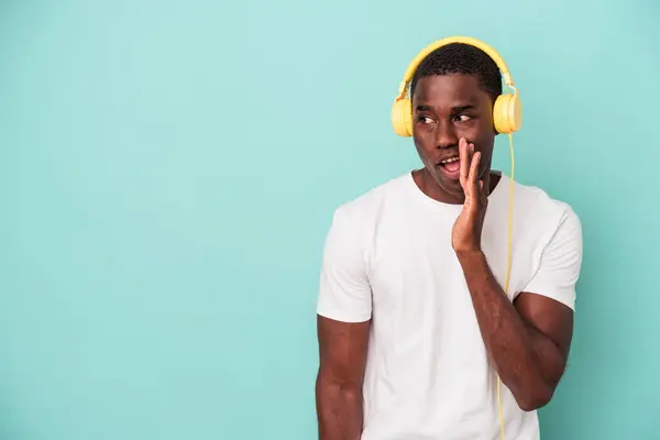 青い背景に隔離された音楽を聞いている若いアフリカ系アメリカ人の男性は 秘密のホットブレーキニュースを言っていると脇を見て — ストック写真
