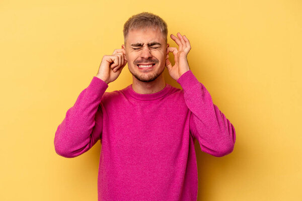 Молодой кавказский мужчина изолирован на желтом фоне, закрывая уши руками.