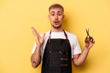 Young caucasian hairdresser man holding scissors isolated on yellow background surprised and shocked.