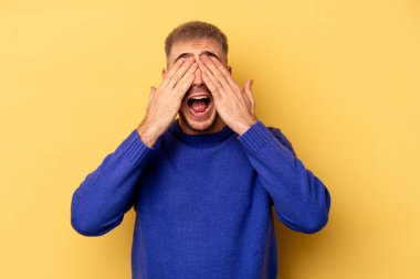 Sarı arka planda izole edilmiş genç beyaz adam elleriyle gözlerini kapatıyor, geniş bir gülümsemeyle bir sürpriz bekliyor..