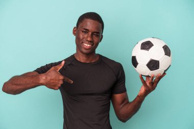 Futbol oynayan genç Afrikalı Amerikalı bir adam mavi arka planda izole edilmiş. Elle bir gömlek fotokopisini işaret ediyor. Gururlu ve kendinden emin.