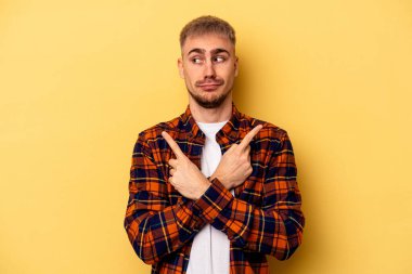 Sarı arka planda izole genç beyaz adam, iki seçenek arasında seçim yapmaya çalışıyor.