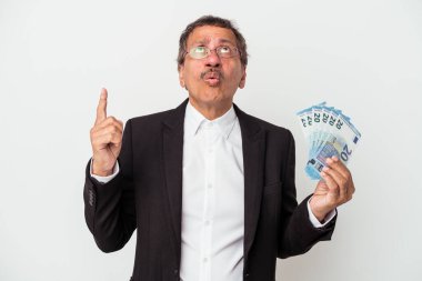 Middle aged indian business man holding bills isolated on white background pointing upside with opened mouth.