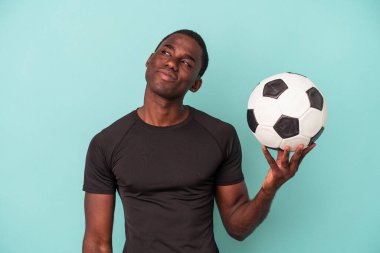 Genç Afrikalı Amerikalı adam mavi arka planda izole bir şekilde futbol oynuyor. Hedeflerine ve amaçlarına ulaşma hayali kuruyor.