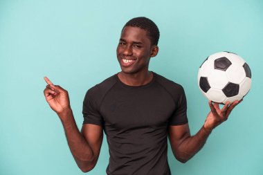 Mavi arka planda izole bir şekilde futbol oynayan genç Afrikalı Amerikalı adam gülümsüyor ve boş alanda bir şeyler gösteriyor..
