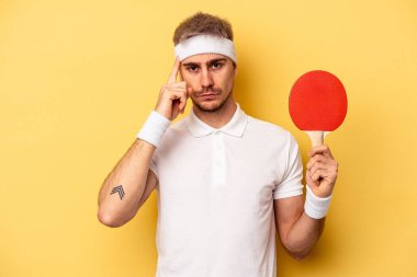 Young caucasian man holding ping pong rackets isolated on yellow background pointing temple with finger, thinking, focused on a task.