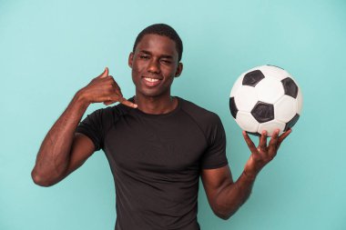 Mavi arka planda izole bir şekilde futbol oynayan genç Afrikalı Amerikalı bir adam parmaklarıyla bir cep telefonu hareketi gösteriyor..