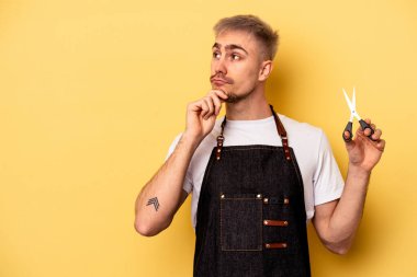 Sarı arka planda izole edilmiş makas tutan Kafkasyalı genç kuaför şüpheli ve şüpheci bir ifadeyle yana bakıyor..