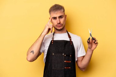 Young caucasian hairdresser man holding scissors isolated on yellow background pointing temple with finger, thinking, focused on a task.