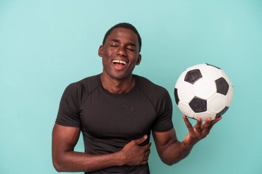 Genç Afro-Amerikan futbol oynayan adam mavi arka planda izole edilmiş gülüyor ve eğleniyor..