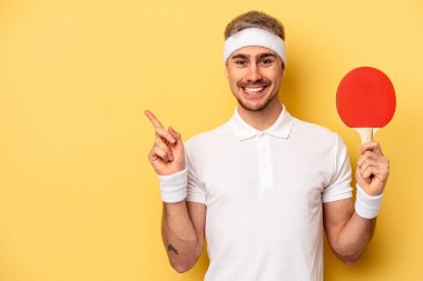 Young caucasian man holding ping pong rackets isolated on yellow background smiling and pointing aside, showing something at blank space.