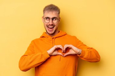 Sarı arka planda izole edilmiş genç beyaz adam gülümsüyor ve elleriyle kalp şeklini gösteriyor..