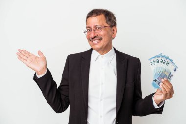 Middle aged indian business man holding bills isolated on white background showing a copy space on a palm and holding another hand on waist.