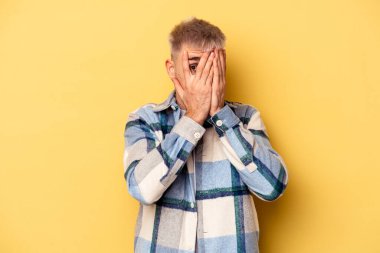 Sarı arka planda izole edilmiş genç beyaz adam parmaklarının arasından korkmuş ve gergin bir şekilde göz kırp..