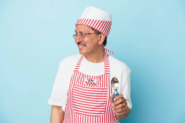 Senior indian ice cream man holding a scoop isolated on blue background looks aside smiling, cheerful and pleasant.