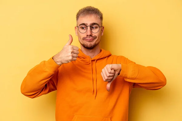 Young caucasian man isolated on yellow background showing thumbs up and thumbs down, difficult choose concept