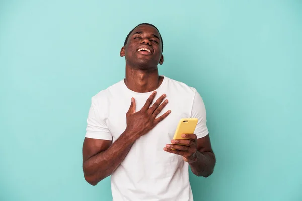 年轻的非裔美国人拿着一部蓝色背景的手机大声笑着把手放在胸前 — 图库照片