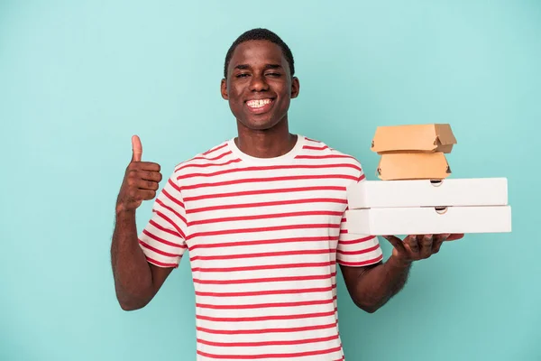 Молодой Афроамериканец Держит Пиццу Бургеры Голубом Фоне Улыбаясь Поднимая Большой — стоковое фото