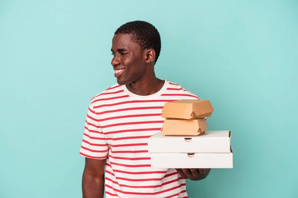 Молодой Афроамериканец Держащий Пиццу Бургеры Голубом Фоне Смотрит Сторону Улыбаясь — стоковое фото