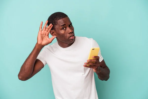 年轻的非裔美国人拿着一部蓝色背景的手机 想听一些闲话 — 图库照片