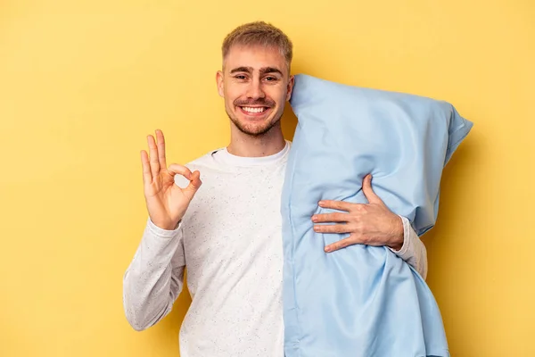 若いですCaucasian男身に着けていますザ Pajamaとホールディング枕隔離上の黄色の背景陽気で自信を持ってショーOkジェスチャー — ストック写真