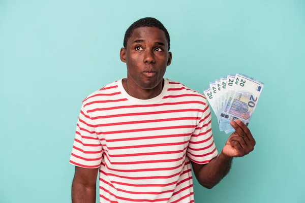 青の背景に隔離された銀行券を保持している若いアフリカ系アメリカ人男性肩と開いて目が混乱 — ストック写真