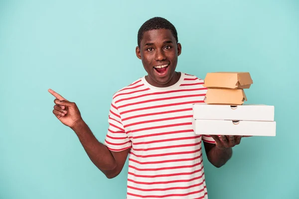 若いアフリカ系アメリカ人の男側を指している青の背景に隔離されたピザやハンバーガーを保持 — ストック写真