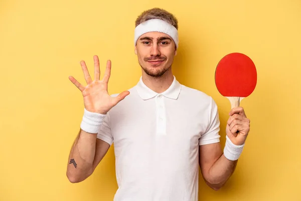若いですCaucasian男保持Ping Pongラケット隔離上の黄色の背景笑顔陽気なショー番号5とともに指 — ストック写真