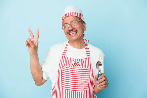 Senior indian ice cream man holding a scoop isolated on blue background joyful and carefree showing a peace symbol with fingers.