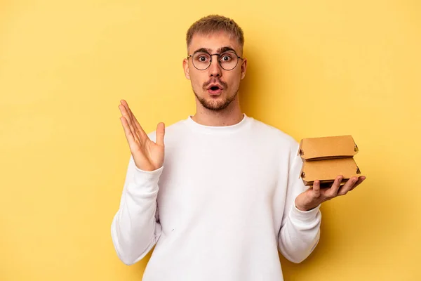 Young Caucasian Man Holding Burger Isolated Yellow Background Surprised Shocked — ストック写真