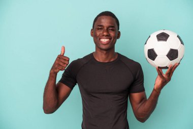 Mavi arka planda izole bir şekilde futbol oynayan genç Afrikalı Amerikalı adam gülümsüyor ve başparmağını kaldırıyor.