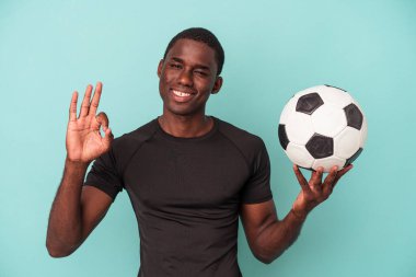 Futbol oynayan genç Afrikalı Amerikalı adam mavi arka planda izole edilmiş neşeli ve kendinden emin bir şekilde iyi bir jest yapıyor..