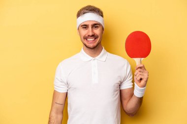 Young caucasian man holding ping pong rackets isolated on yellow background happy, smiling and cheerful.