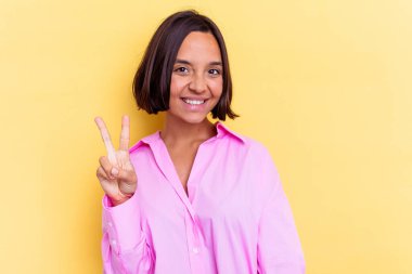 Sarı arka planda izole edilmiş genç melez kadın zafer işareti gösteriyor ve gülümsüyor..