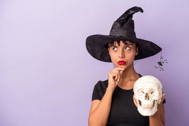 Mor arka planda kafatasını tutan cadı kılığına girmiş genç bir ırk kadını şüpheli ve şüpheci bir ifadeyle yan yana bakıyor..