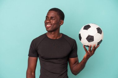 Mavi arka planda izole bir şekilde futbol oynayan genç Afrikalı Amerikalı adam gülümsüyor, neşeli ve keyifli görünüyor..
