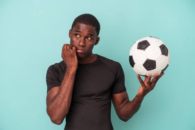Genç Afrikalı Amerikalı bir adam futbol oynuyor. Mavi arka planda izole edilmiş, tırnaklarını yiyor. Gergin ve endişeli..
