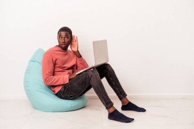 Genç Afrikalı Amerikalı bir adam beyaz arka planda izole edilmiş bir dizüstü bilgisayar kullanarak bir dedikoduyu dinlemeye çalışıyor..