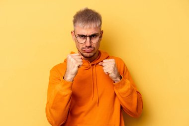 Sarı arka planda izole edilmiş genç beyaz adam kameraya yumruk gösteriyor, agresif yüz ifadesi var..